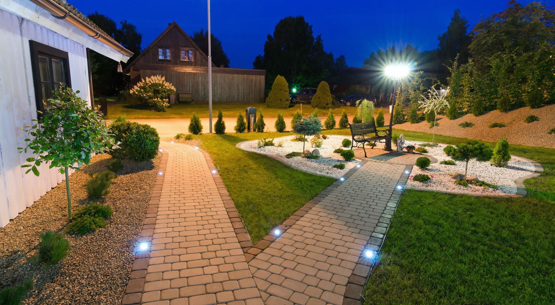 Night view of a landscaped yard with illuminated brick pathways leading to a bench and garden features.