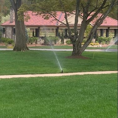 Lush green lawn with sprinklers watering, in front of a house with a red-tiled roof.