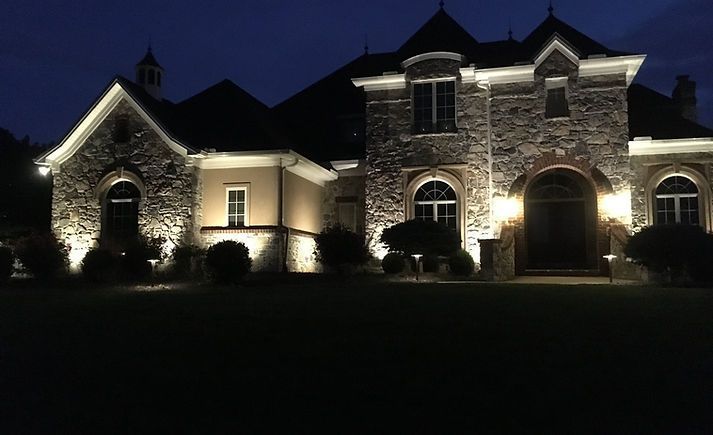 Night view of a stone house lit up with landscape lighting. Dark blue sky.