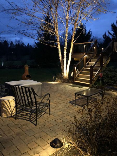 Brick patio with outdoor lighting around a tree, stairs, and furniture at dusk.