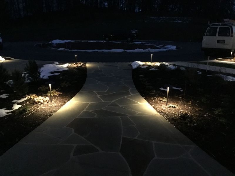 Lit pathway at night, featuring stone paving and outdoor lights, with patches of snow.