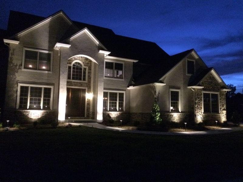 Two-story house illuminated at night with spotlights and landscaping lights; blue sky backdrop.