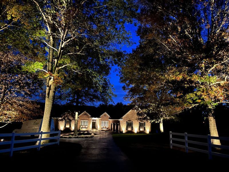 Illuminated house at night with trees and a white fence under a dark blue sky.