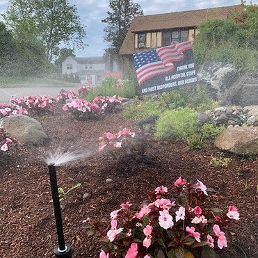 Sprinkler watering flowers in a landscaped yard; sign thanks healthcare workers with American flags.
