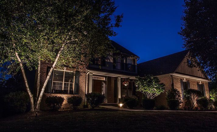 Illuminated two-story brick house at night; landscape lighting highlights trees and facade under a dark blue sky.