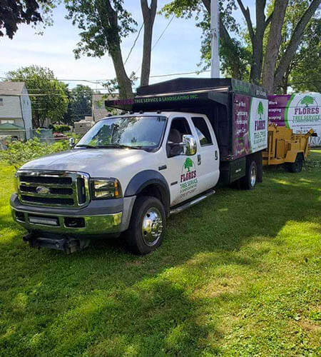 White tree service truck with a black dump bed, and a yellow wood chipper on a grassy lawn.