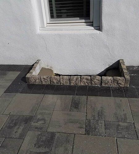 A rectangular planter made of decorative blocks sits below a window on a white wall.