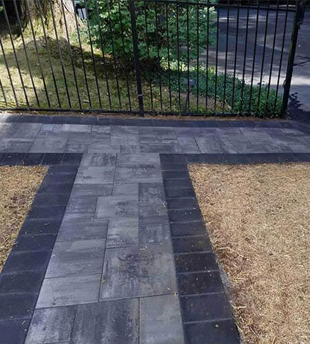 A brick pathway in front of a black fence, with brown grass and a small tree.