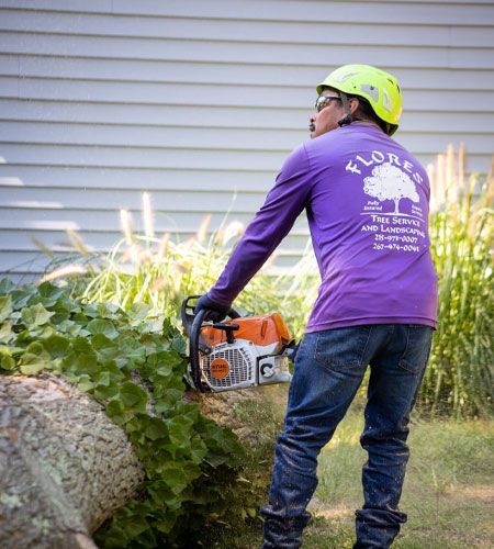 Person in safety gear using a chainsaw to cut a log near a building.