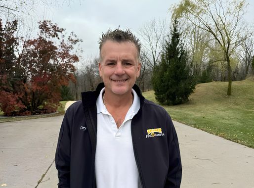 Man smiling outdoors, wearing a black jacket over a white shirt. Green trees and a path in background.