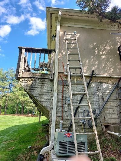 Ladders leaning against a house exterior beside a small deck, with a gutter downspout and grassy yard.