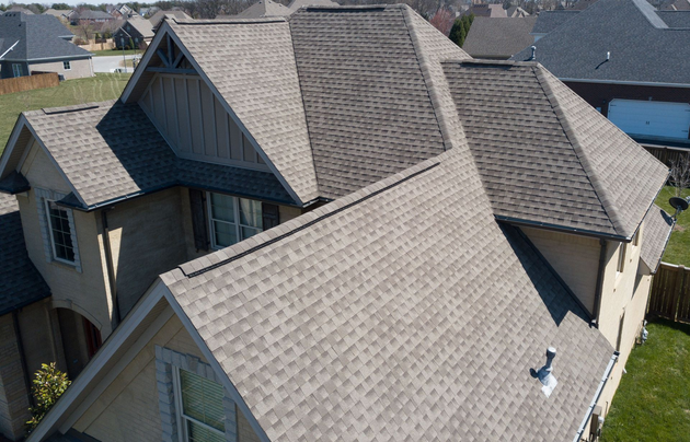 A close-up of a house roof with green shingles, multiple gables, dormer windows, and light-colored wooden siding.