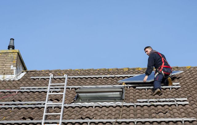 A worker wearing a safety harness installs a solar panel on a tiled roof under a clear blue sky.