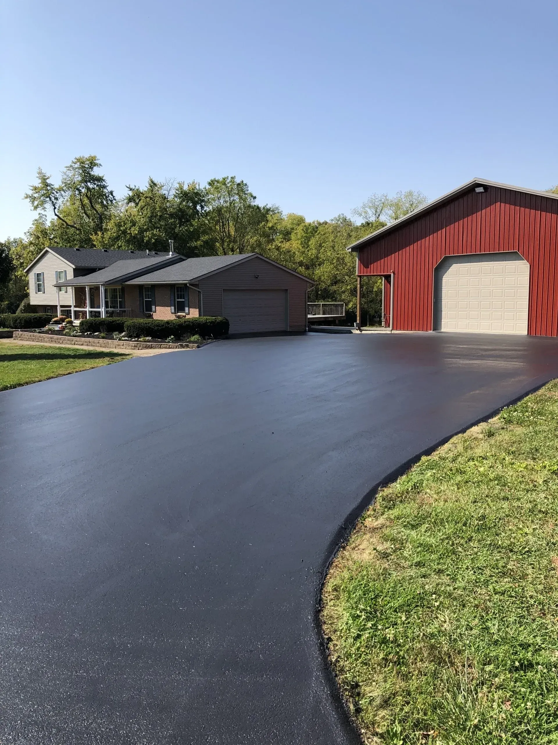 A newly paved asphalt driveway leads to a suburban house and a large red barn under a clear blue sky.
