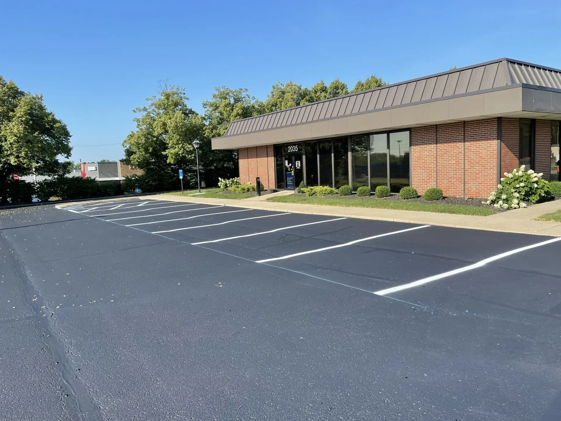 A freshly paved asphalt parking lot with white-lined spaces in front of a one-story brick commercial building under a sky.
