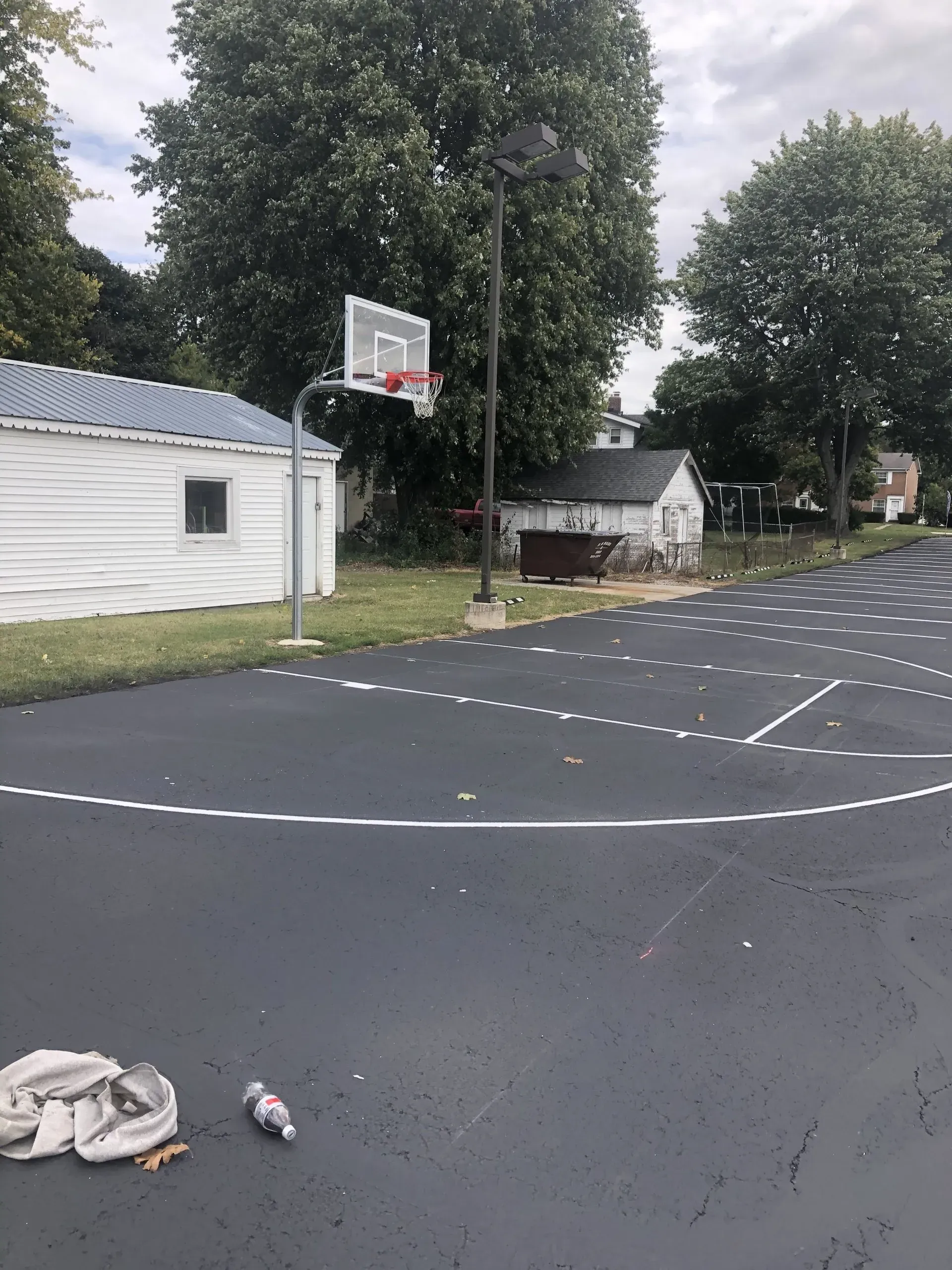 A basketball court with white markings on dark pavement, a hoop, white buildings, and trees under a cloudy sky.