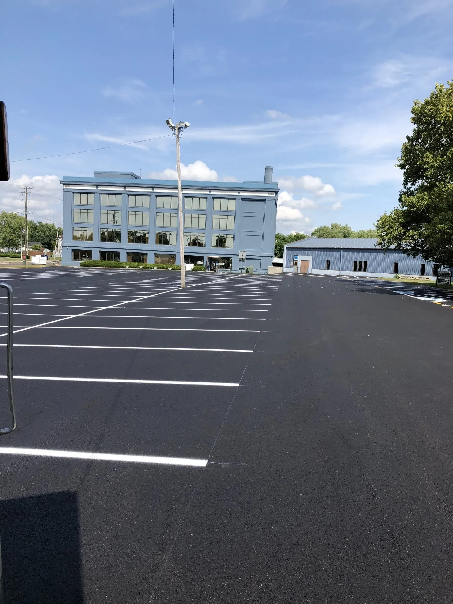 A newly paved parking lot with white painted spaces in front of a modern blue office building under a clear blue sky.