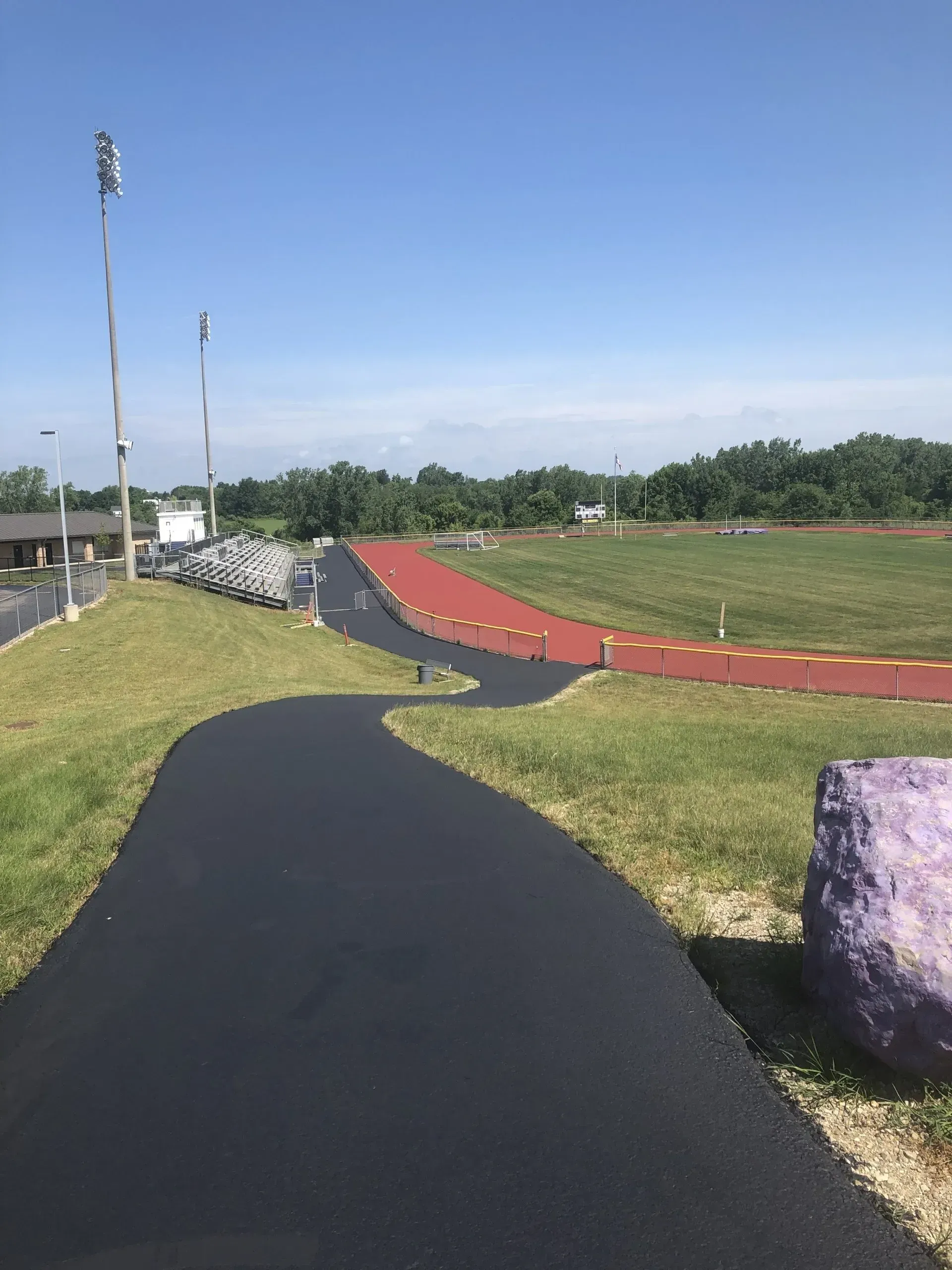 A black asphalt path curves toward a red athletic track surrounding a grassy field under a clear blue sky.