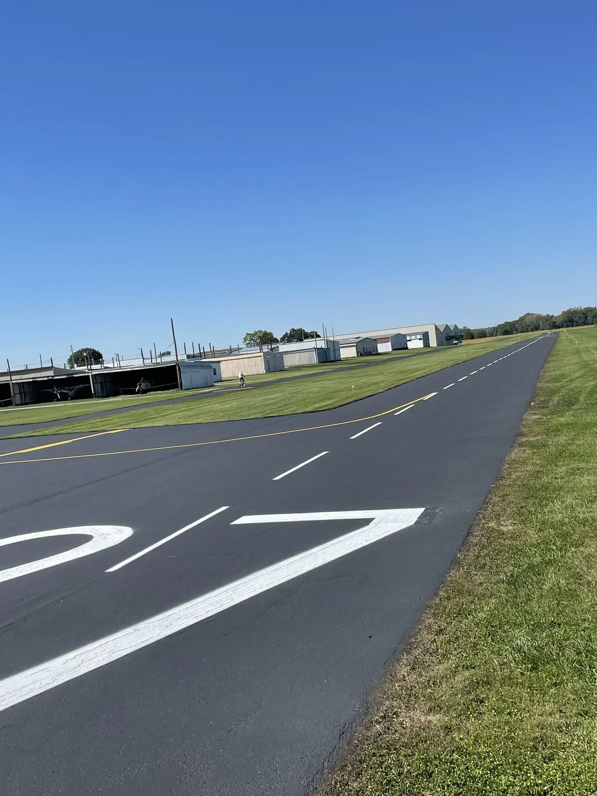 A freshly paved runway or taxiway with white markings, adjacent to a grassy field and a line of buildings under a blue sky.