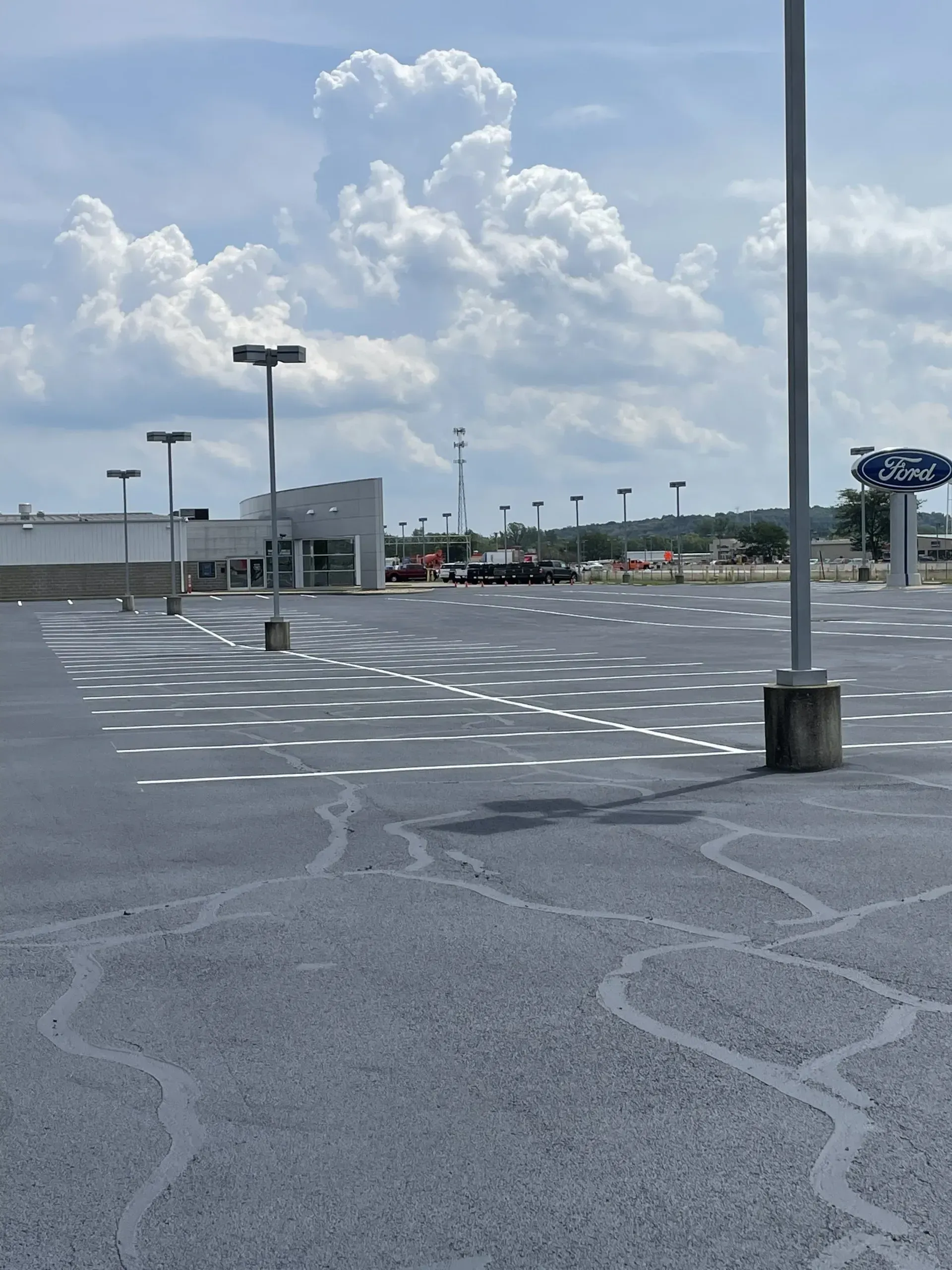 An empty Ford dealership parking lot under a cloudy blue sky with light poles and faded white parking space markings.
