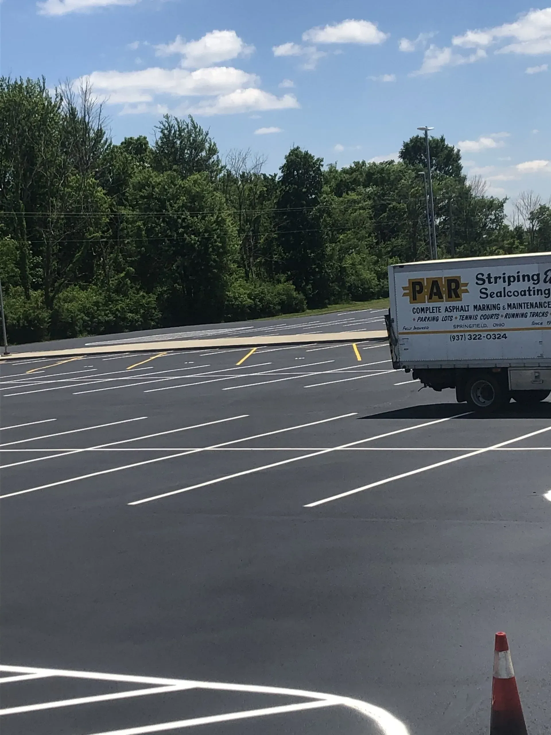 A striping truck in a newly paved parking lot with white paint markings and a traffic cone in the foreground.