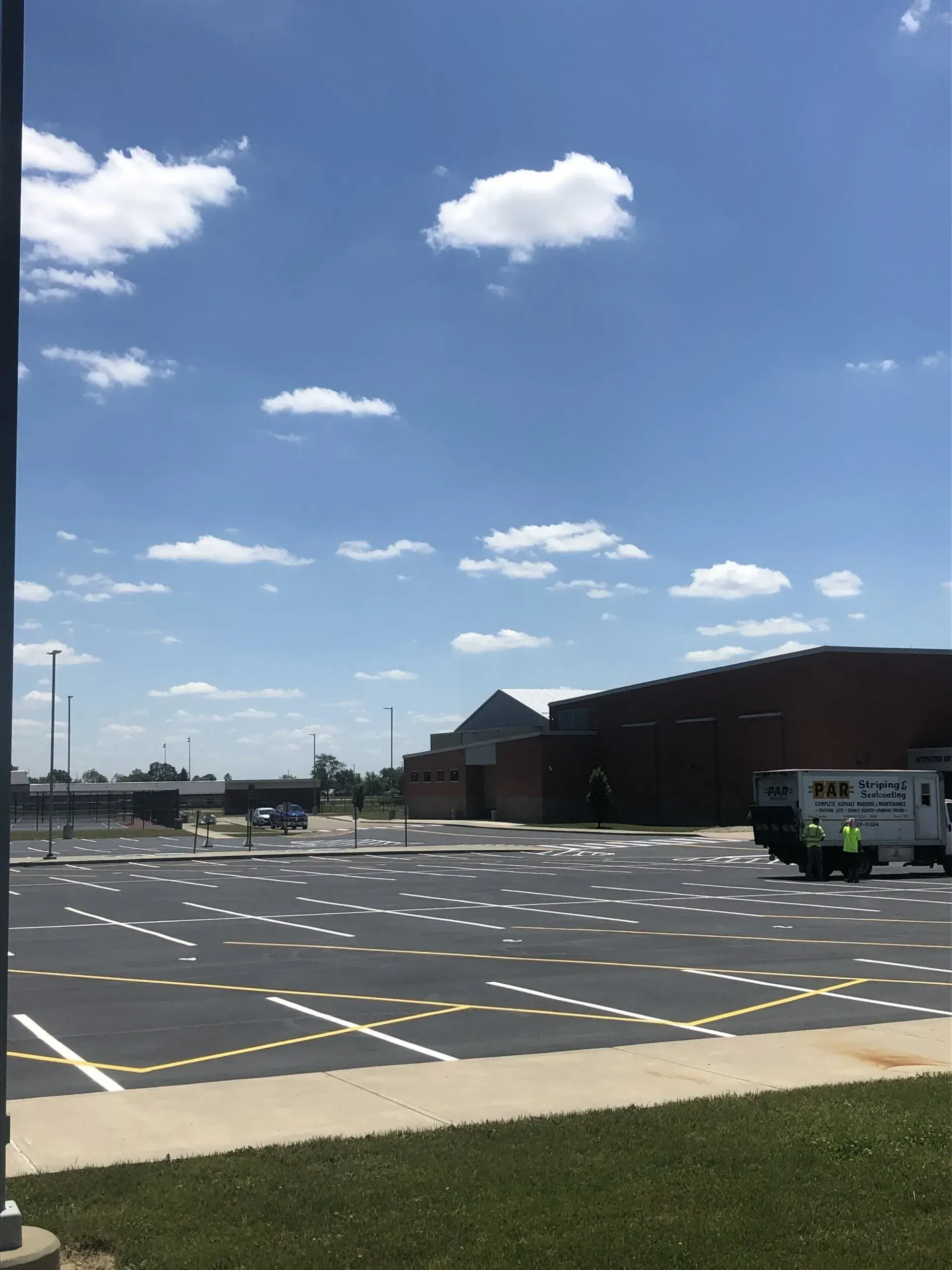 A parking lot marked with white grids under a sunny, blue sky with a brick building and a utility vehicle in the background.