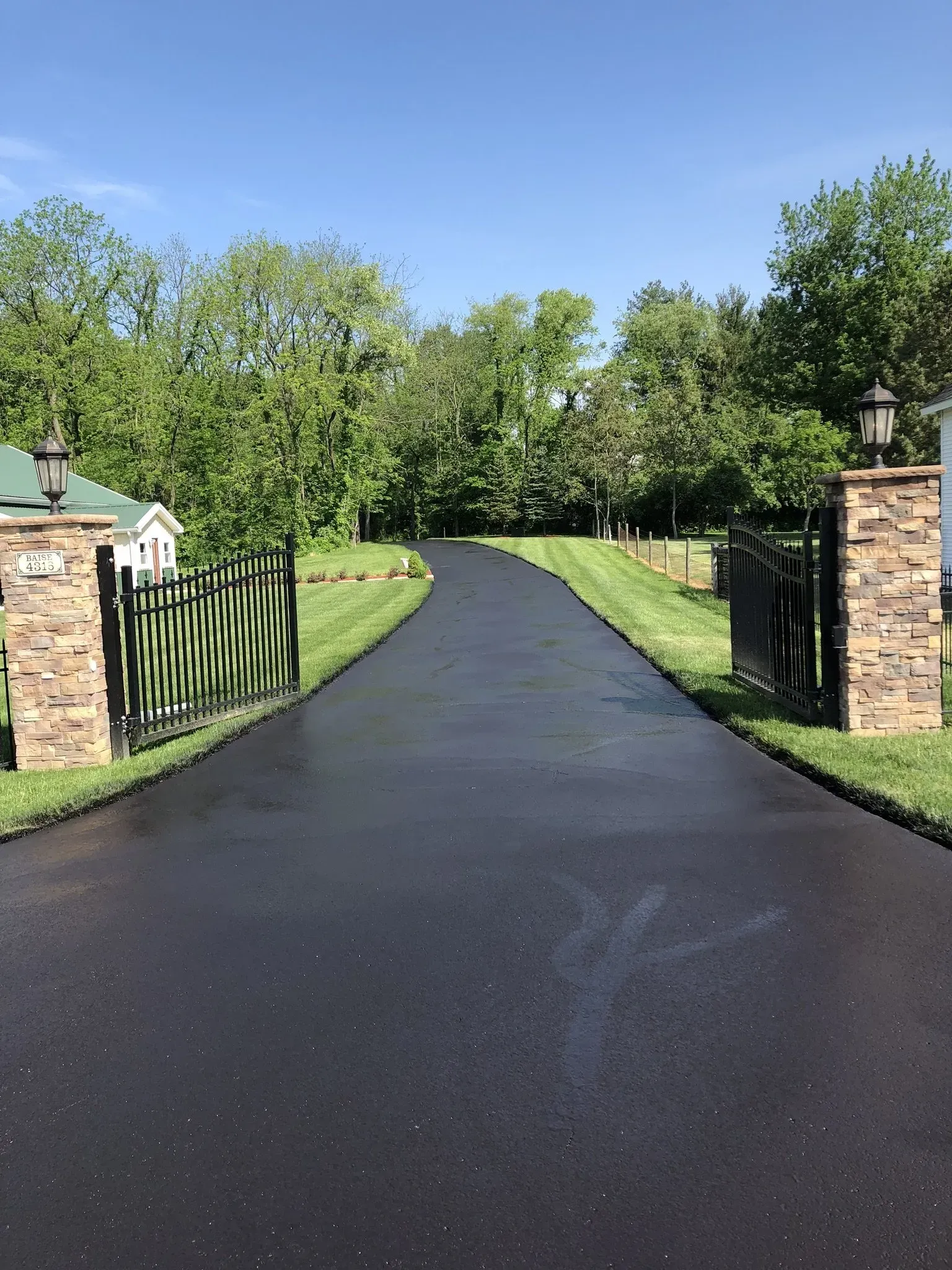 A black asphalt driveway curves between two stone pillars topped with lanterns, bordered by grass and green trees.