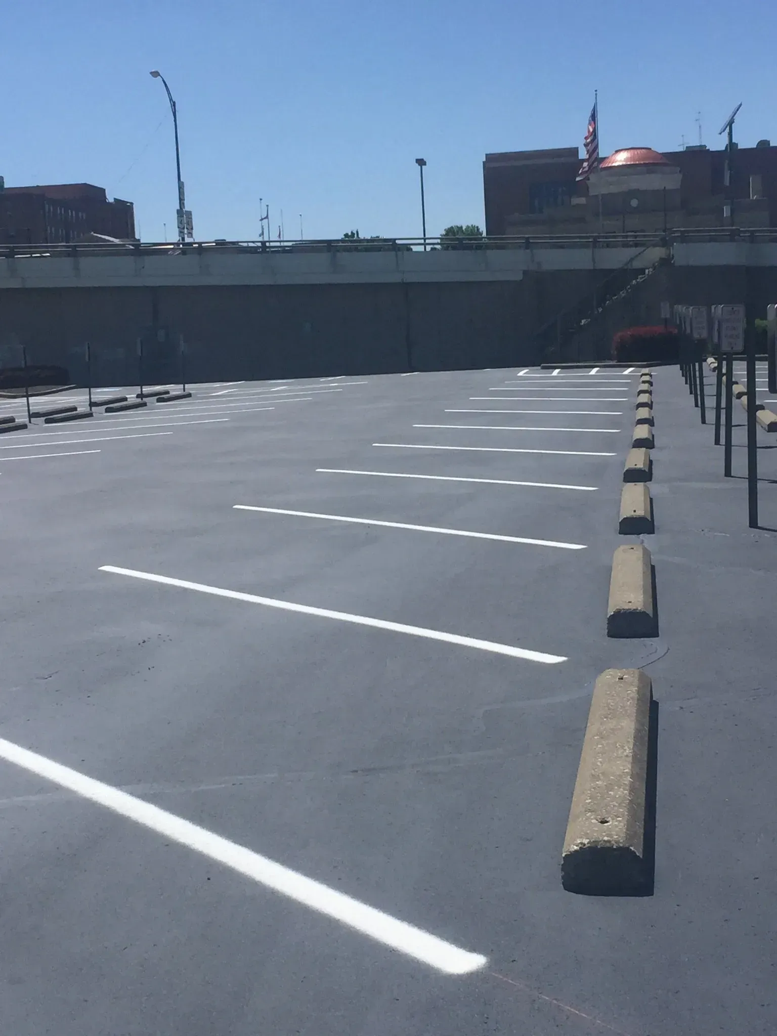 Empty asphalt parking lot with white painted stall lines and concrete wheel stops under a clear blue sky.