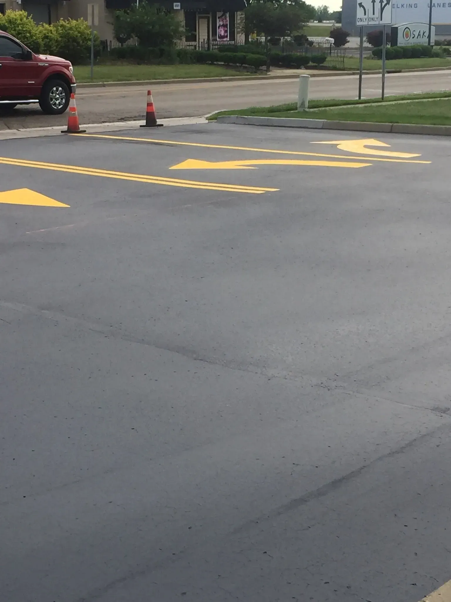 An asphalt parking lot entrance featuring yellow painted directional arrows and dashed lines with two orange traffic cones.