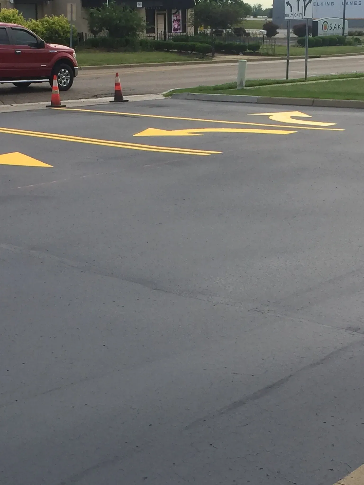A parking lot exit with yellow painted directional arrows, a dashed stop line, and two traffic cones.