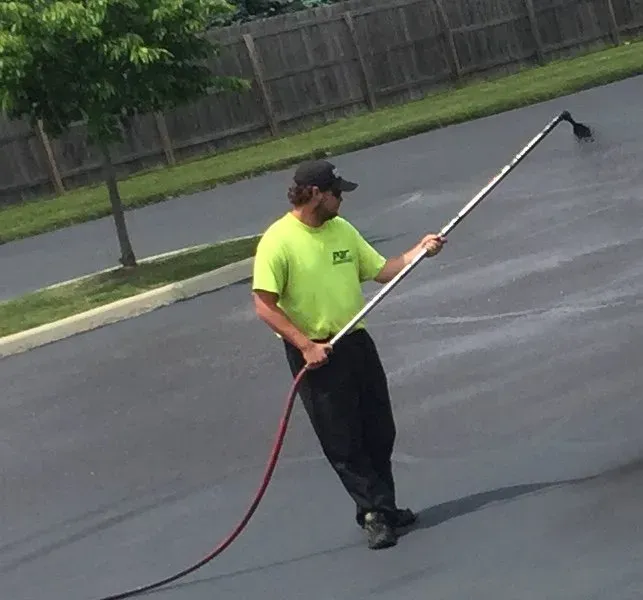 A person in a neon yellow shirt uses a long-handled sprayer to apply sealant to a grey asphalt parking lot.