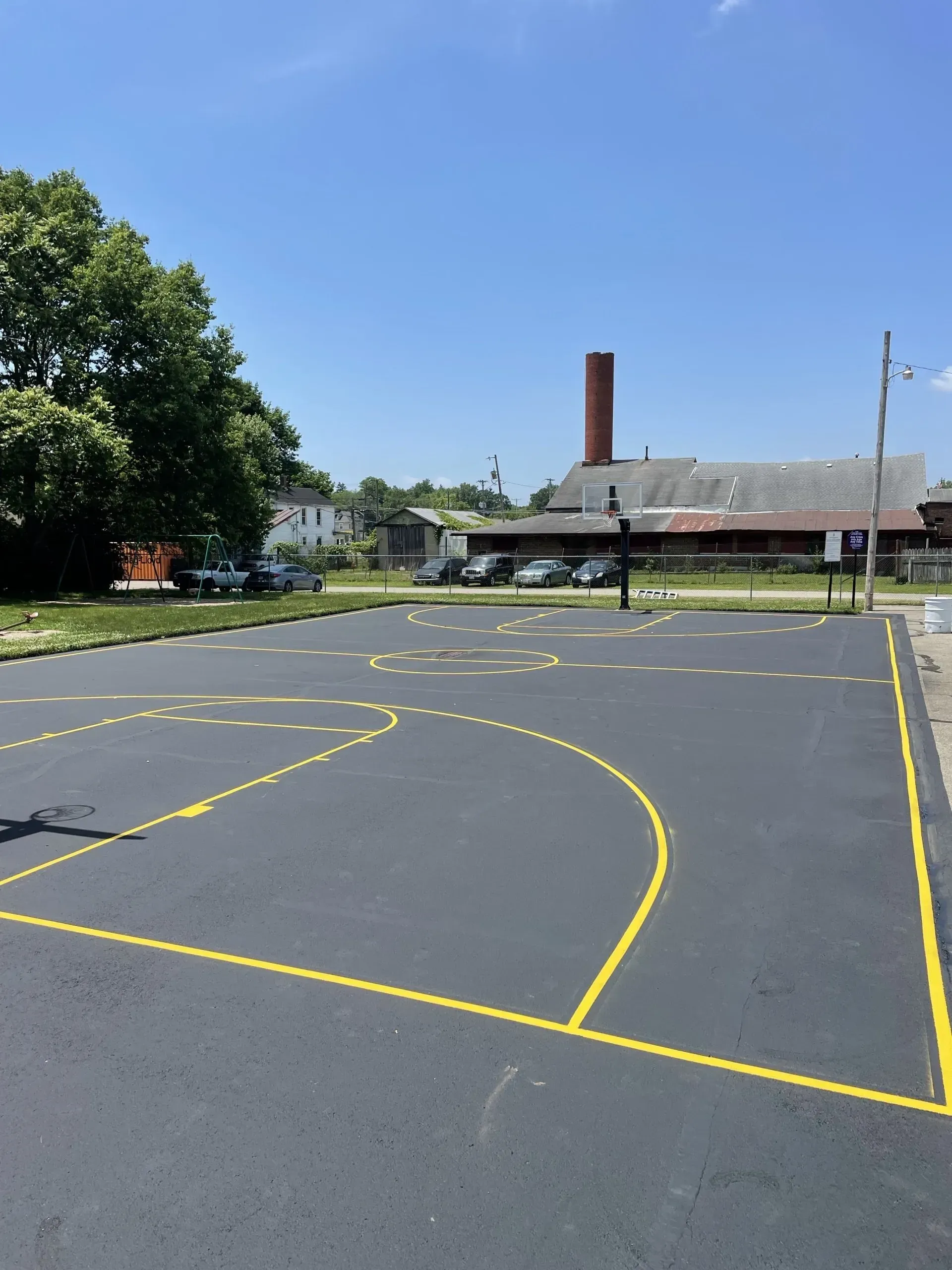 An outdoor basketball court with yellow painted lines on black asphalt, set against a blue sky and industrial buildings.