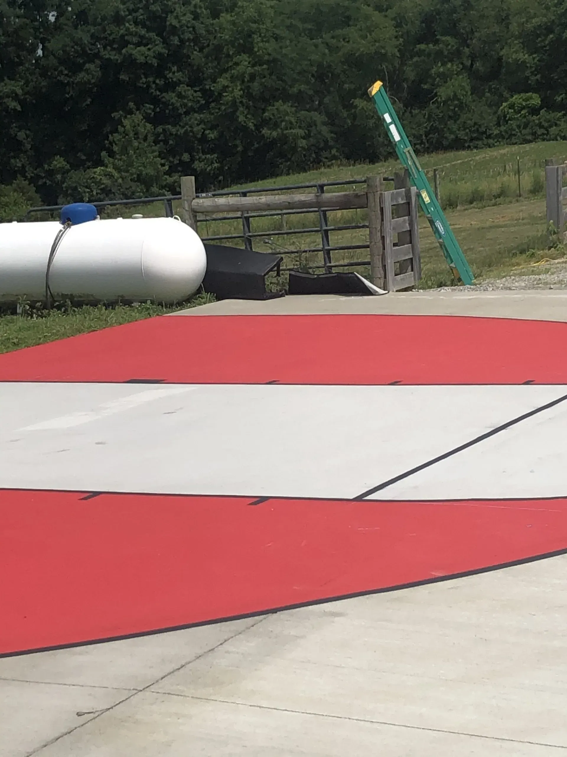 A red and white cover laid out on a concrete surface near a white propane tank and a fence in a grassy area.