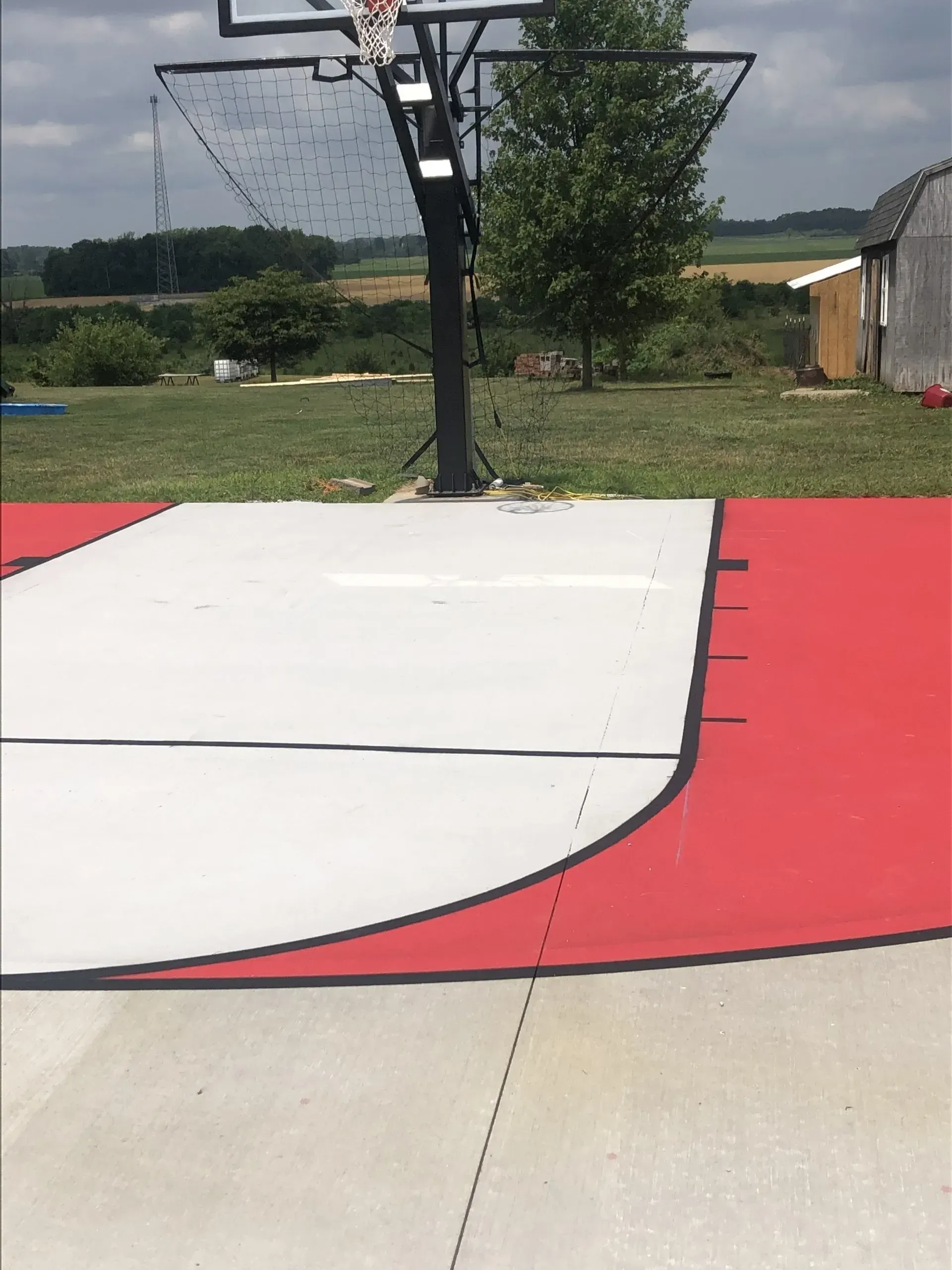 An outdoor basketball court with a grey key, red outer perimeter, and a black hoop pole set against a rural landscape.