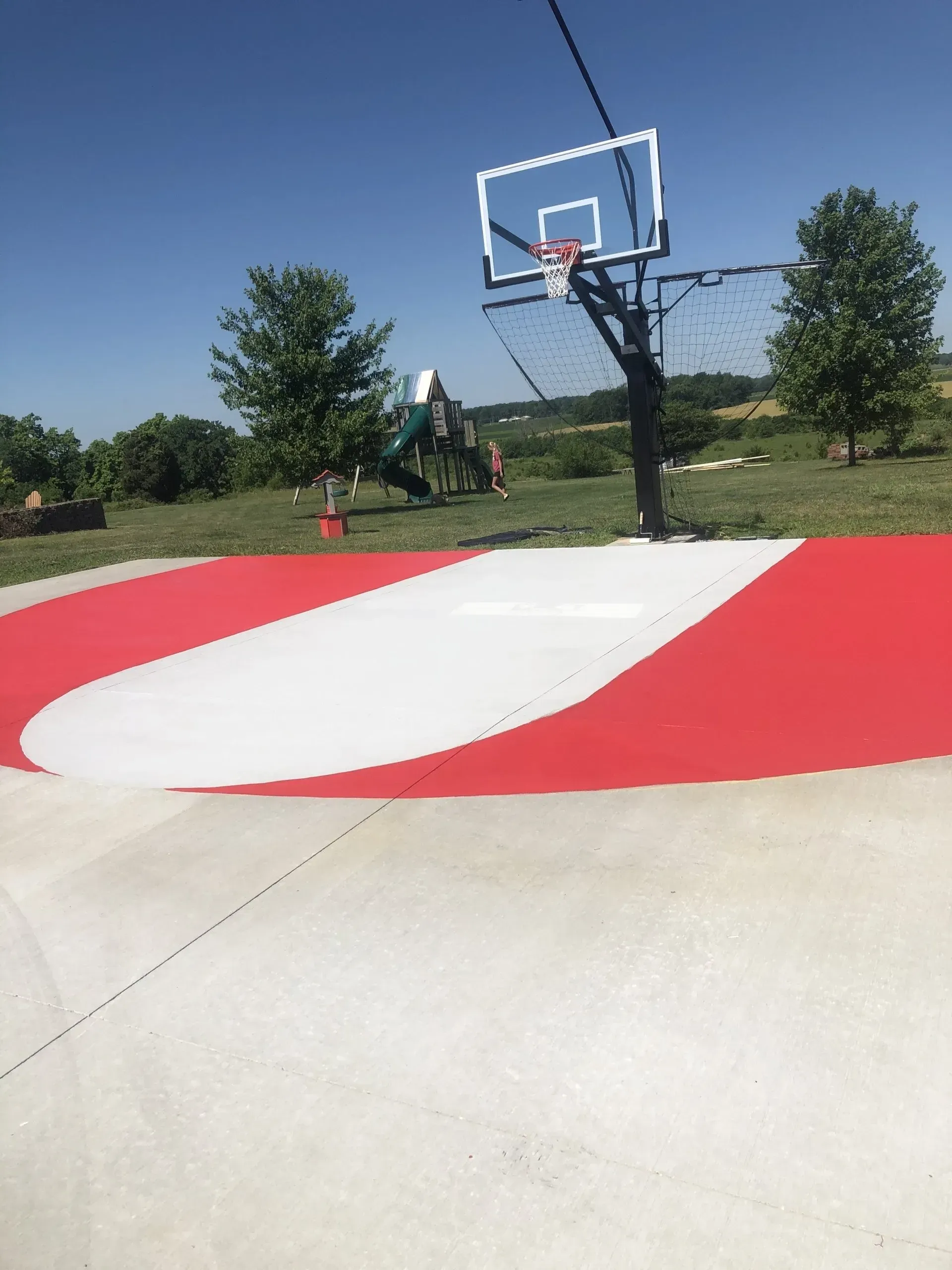 A basketball hoop stands on a driveway with a large, painted white and red court design against a grassy, sunny landscape.