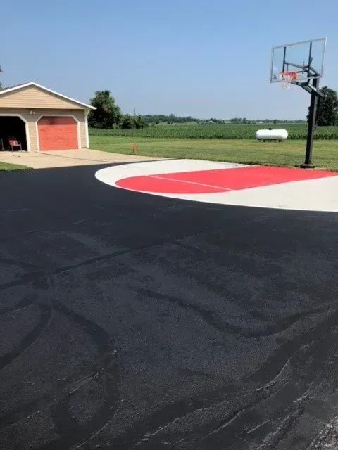 A basketball hoop stands on a driveway with a painted red and white key, next to a detached garage and a grassy field.
