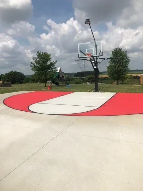 Outdoor basketball hoop on a concrete court with a red and white painted key against a cloudy blue sky.