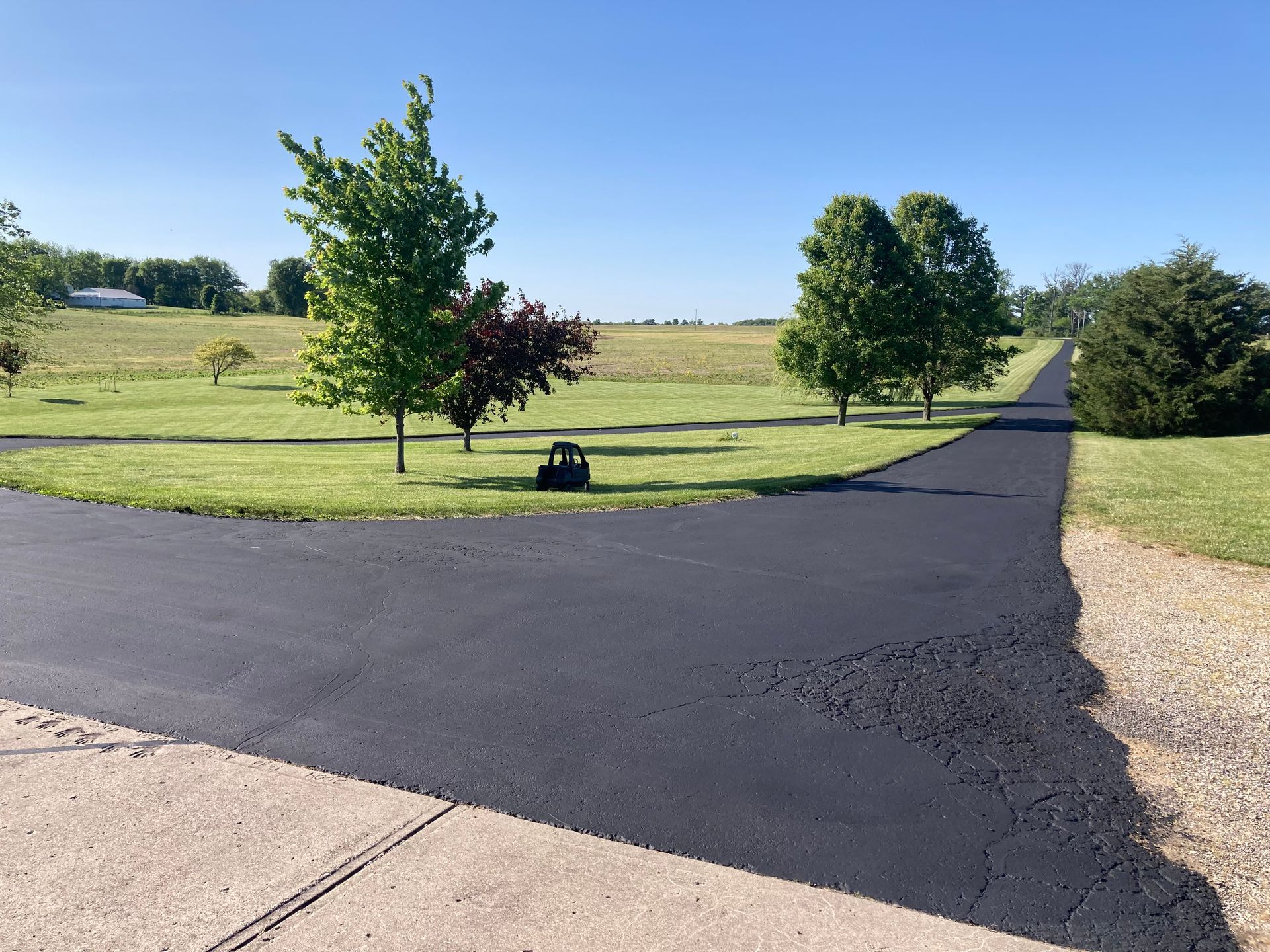 A freshly paved black asphalt driveway curves through a green, sunny rural landscape with several trees.