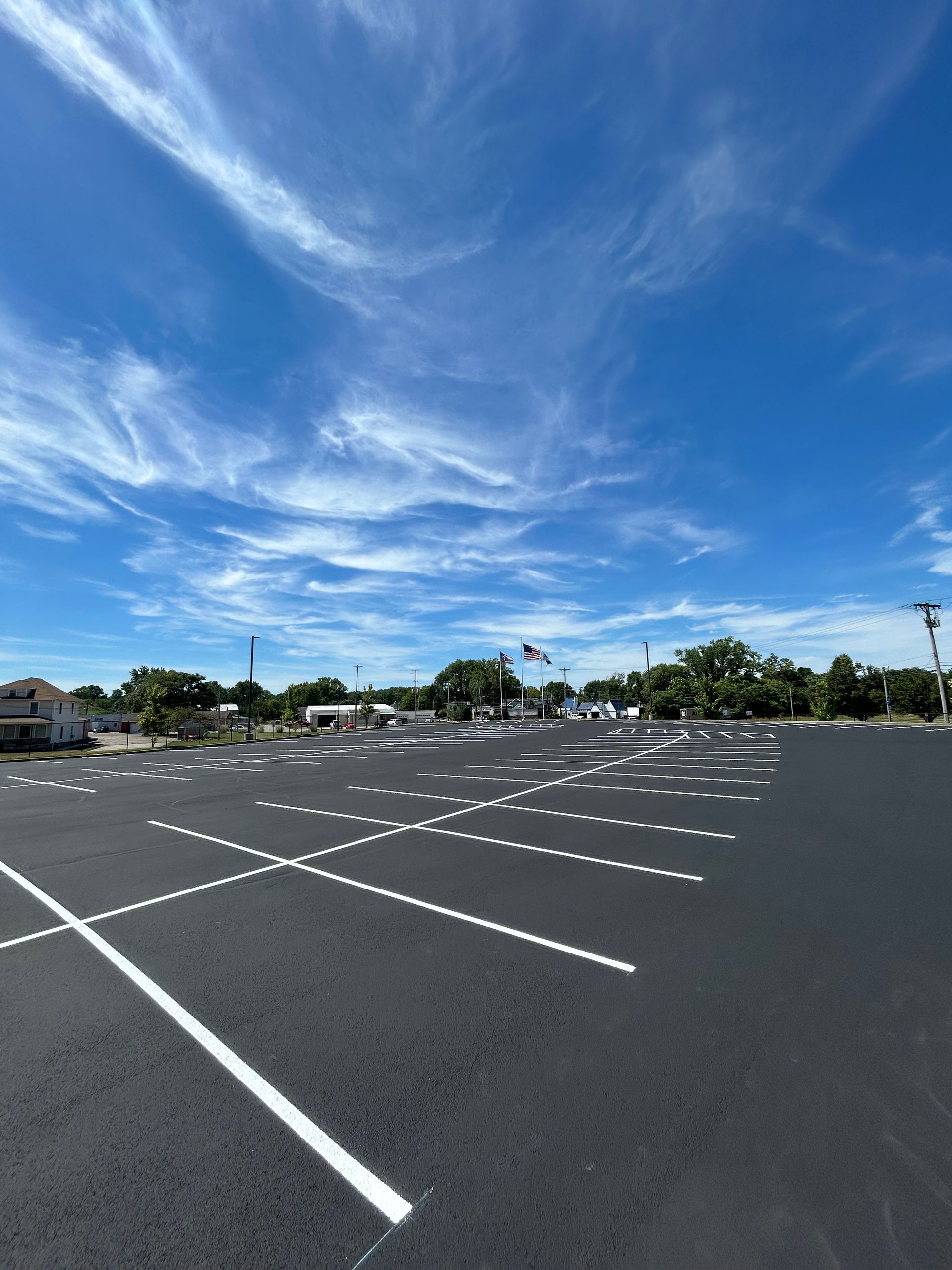 A wide-angle view of a freshly paved, empty parking lot under a bright blue sky with wispy clouds.