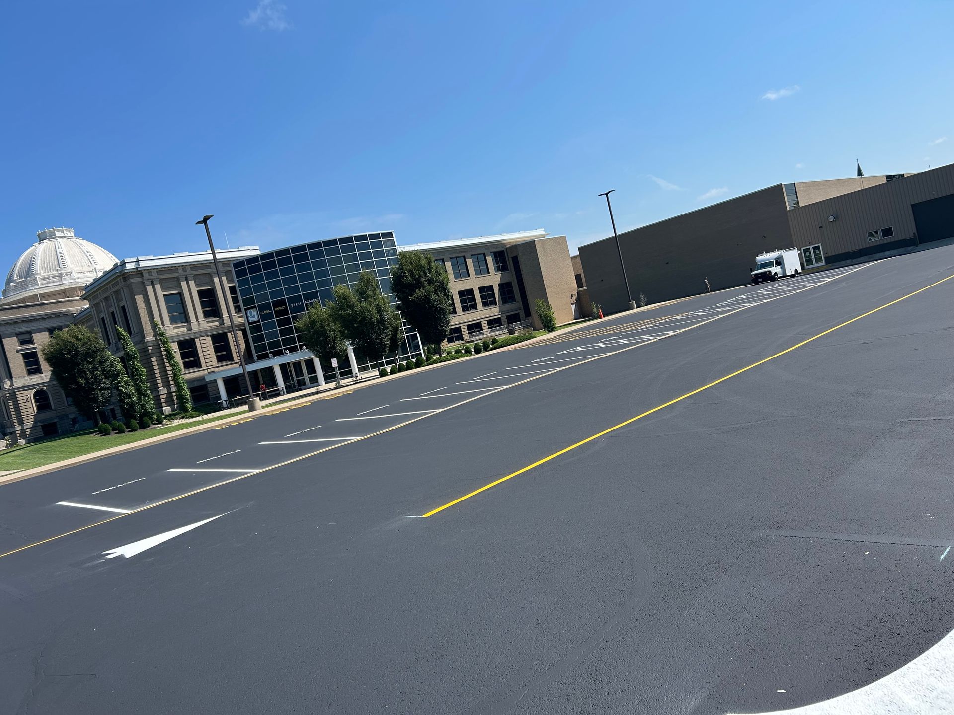 A large, historic-style building with a dome sits behind a wide, empty paved lot under a bright blue sky.