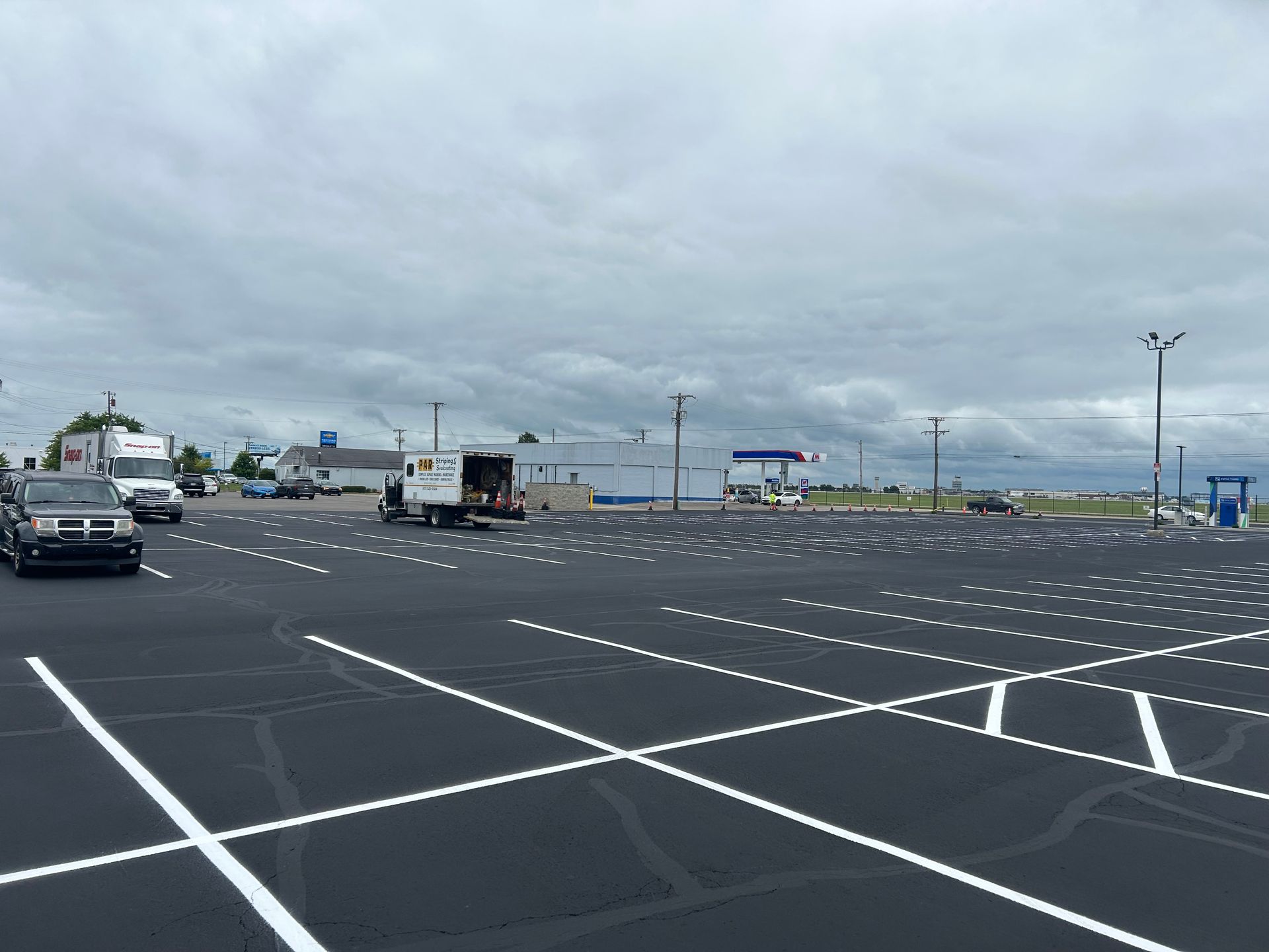 A mostly empty, newly striped asphalt parking lot under a cloudy sky, with a few service vehicles parked in the distance.
