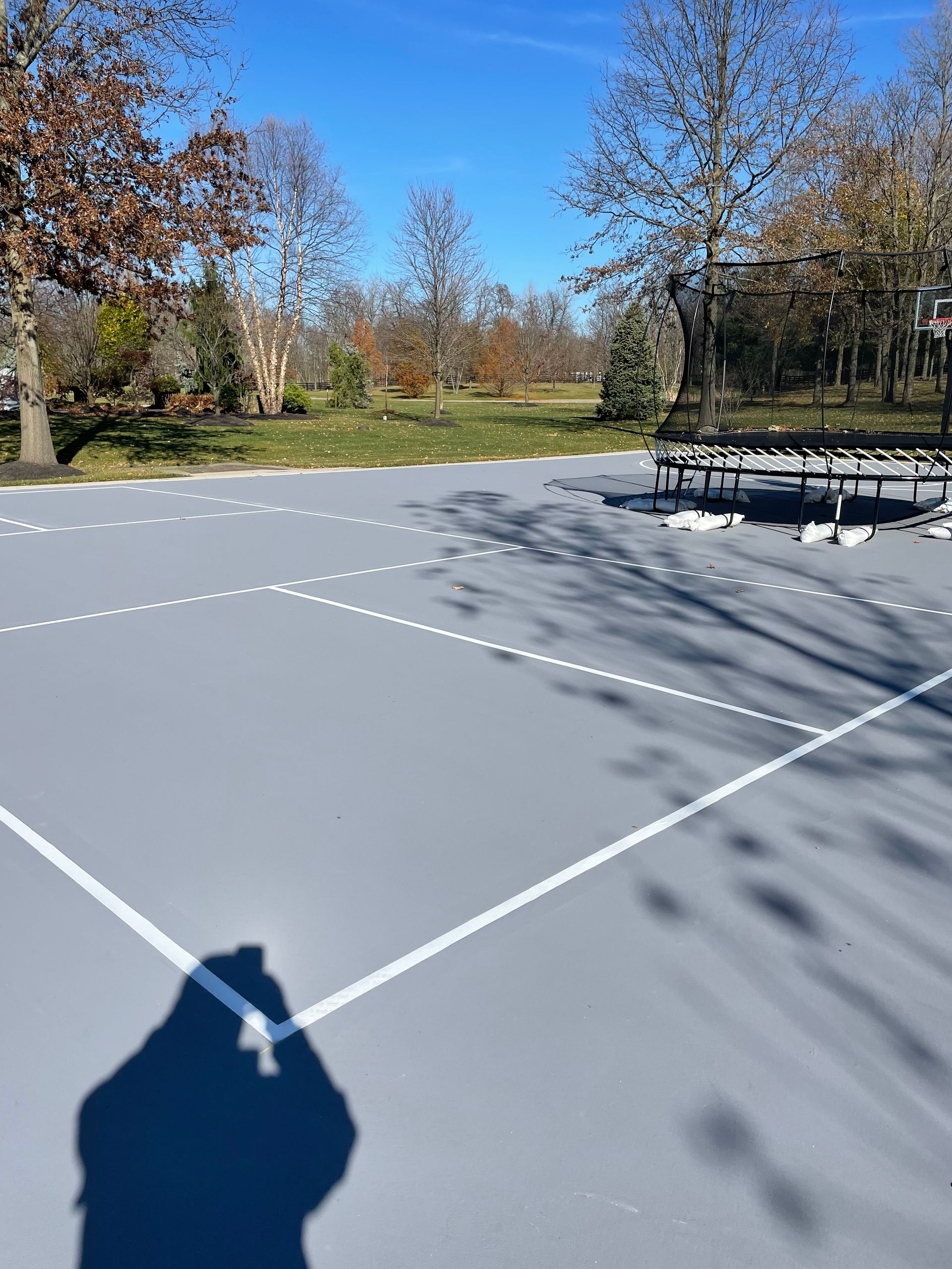 A gray outdoor sports court with white boundary lines, set against a background of trees under a clear blue sky.