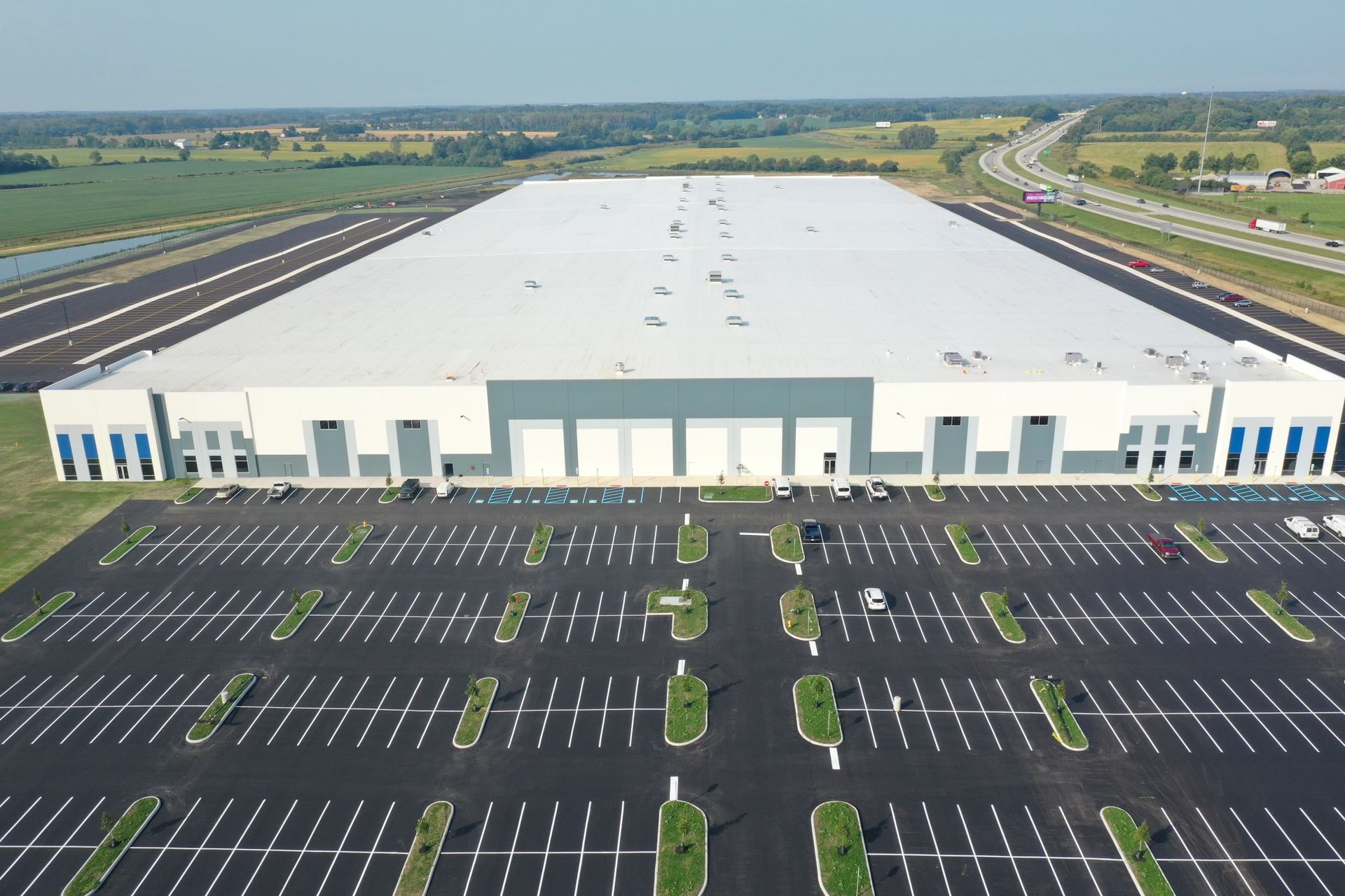 An aerial view of a large white and gray industrial warehouse with a vast parking lot in front, surrounded by greenery.