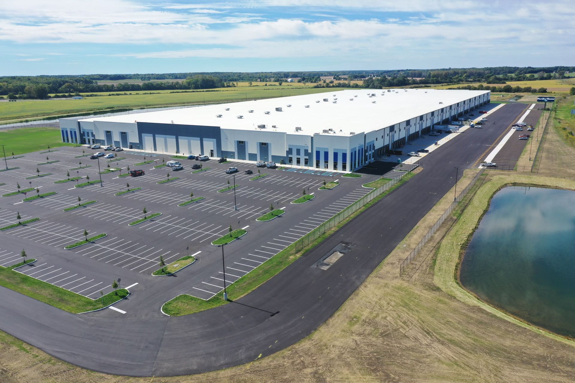 Aerial view of a large white industrial warehouse with a vast parking lot and a retention pond, set in a grassy landscape.