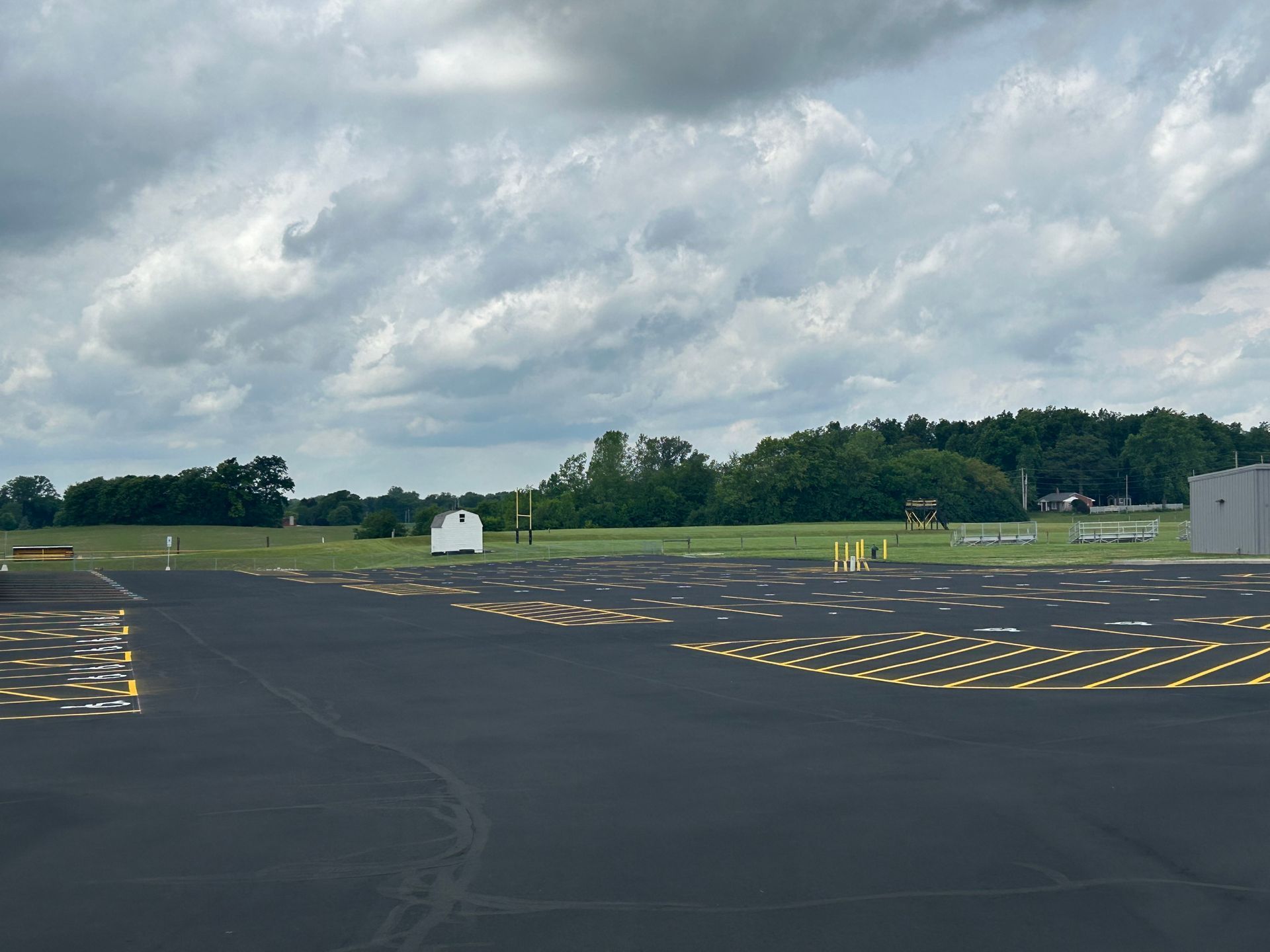 An empty asphalt parking lot with yellow painted markings under a cloudy sky, bordering a grassy field and distant trees.