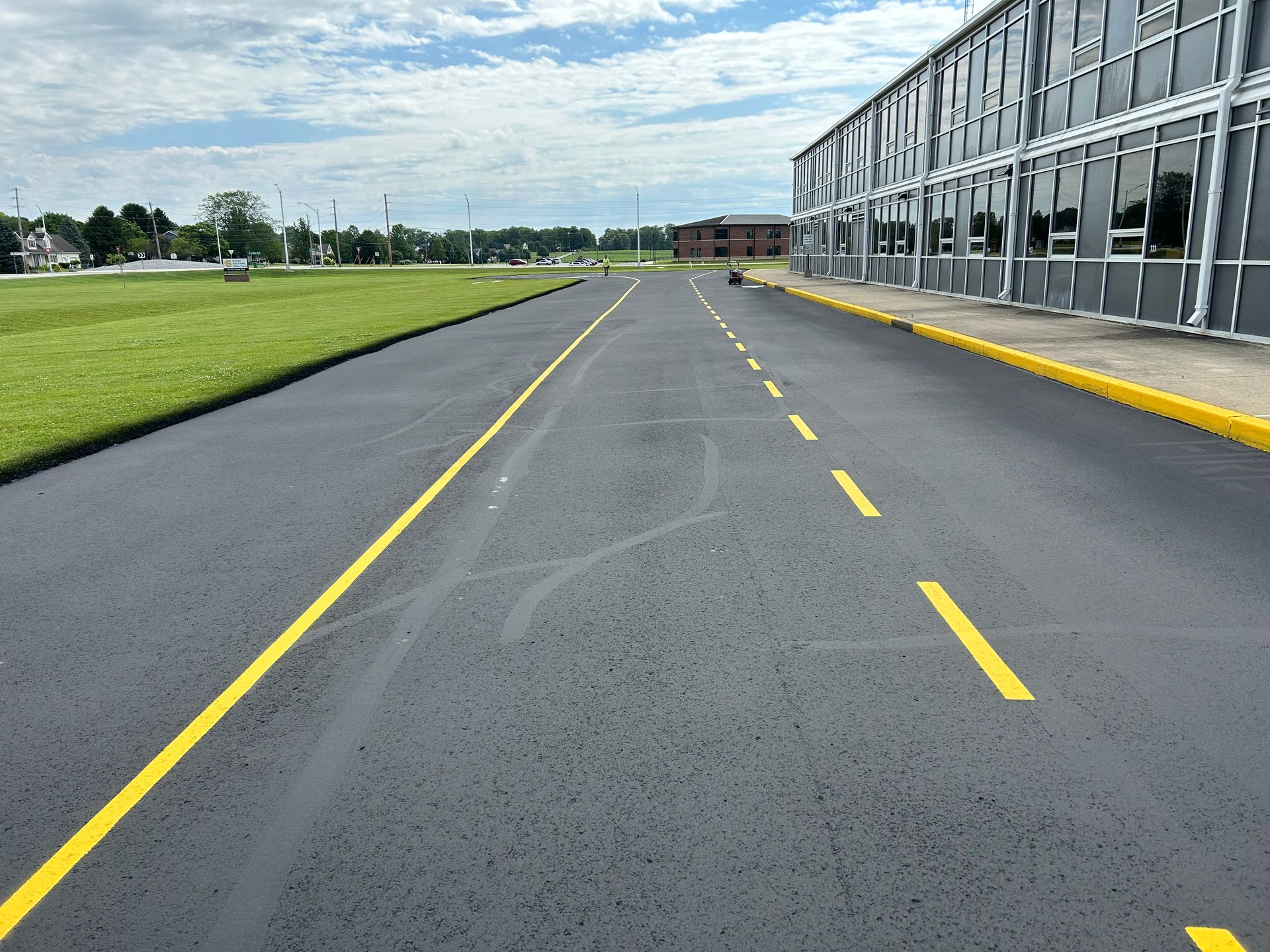 A freshly paved road runs alongside a modern building with yellow painted curbs and lane markings under a cloudy sky.
