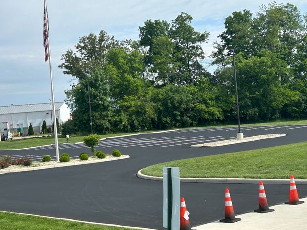 A freshly paved black asphalt parking lot with white marked spaces, orange traffic cones, and a flag pole on a sunny day.