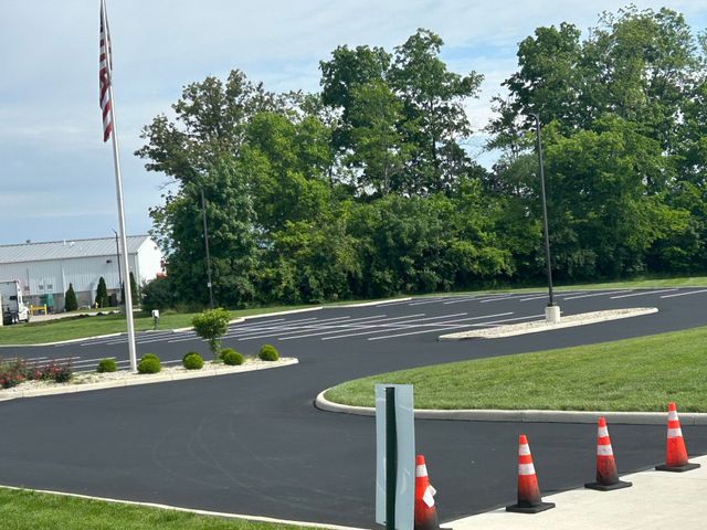 A freshly paved black asphalt parking lot with white marked spaces, orange traffic cones, and a flag pole on a sunny day.