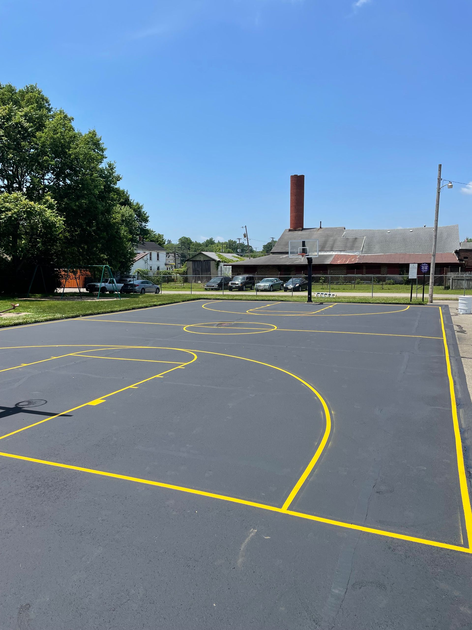 A newly paved basketball court with bright yellow painted lines, set against a background of trees and a brick building.