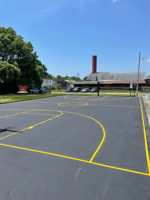A freshly paved outdoor basketball court with bright yellow painted lines, set against a background of trees and buildings.